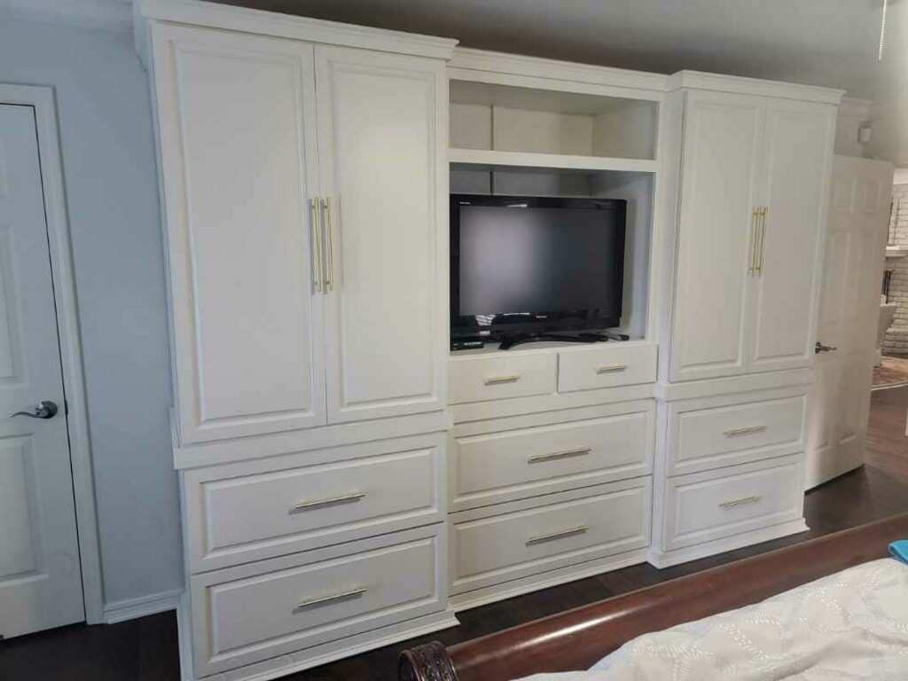 White built-in bedroom cabinet with TV, drawers, and tall storage on a wood floor.