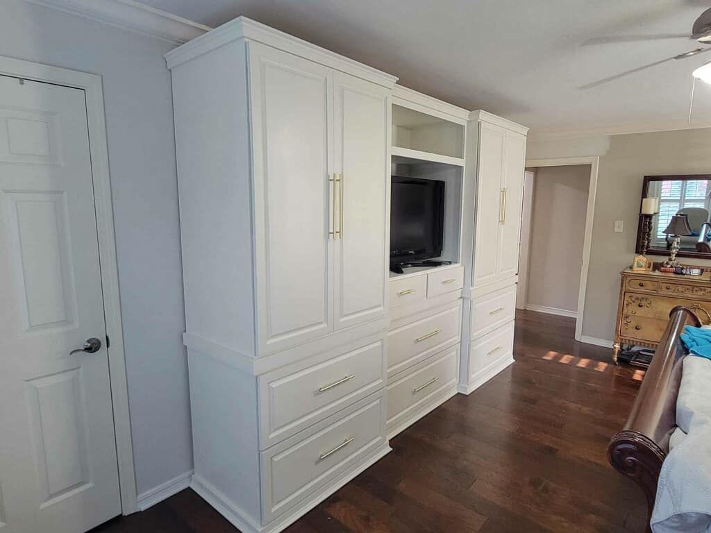 White built-in bedroom cabinetry with doors, drawers, and TV. Brown hardwood floor and white walls.