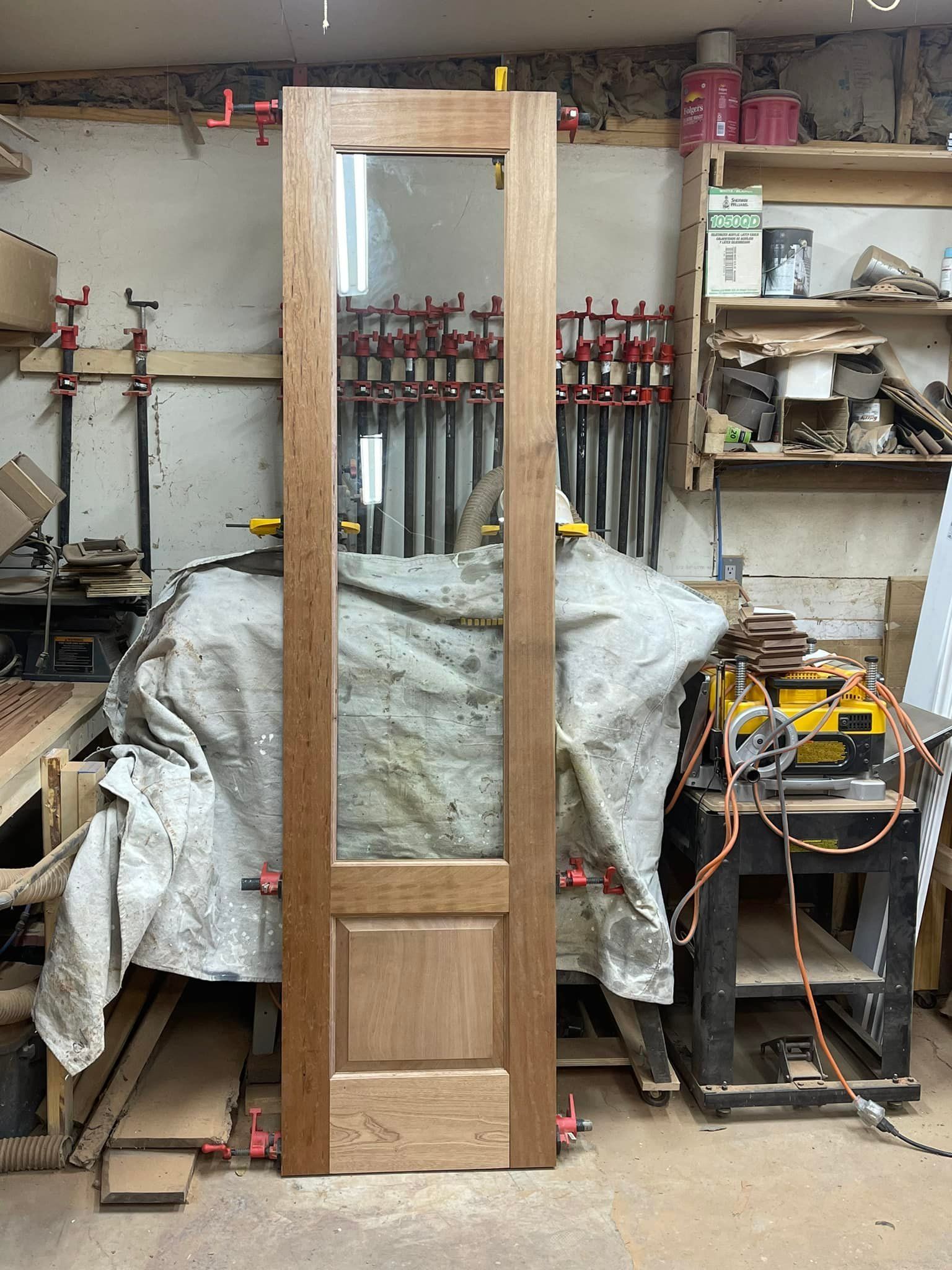 Wooden door with glass panel, clamped in a workshop. Light wood, tools, and a cluttered background.