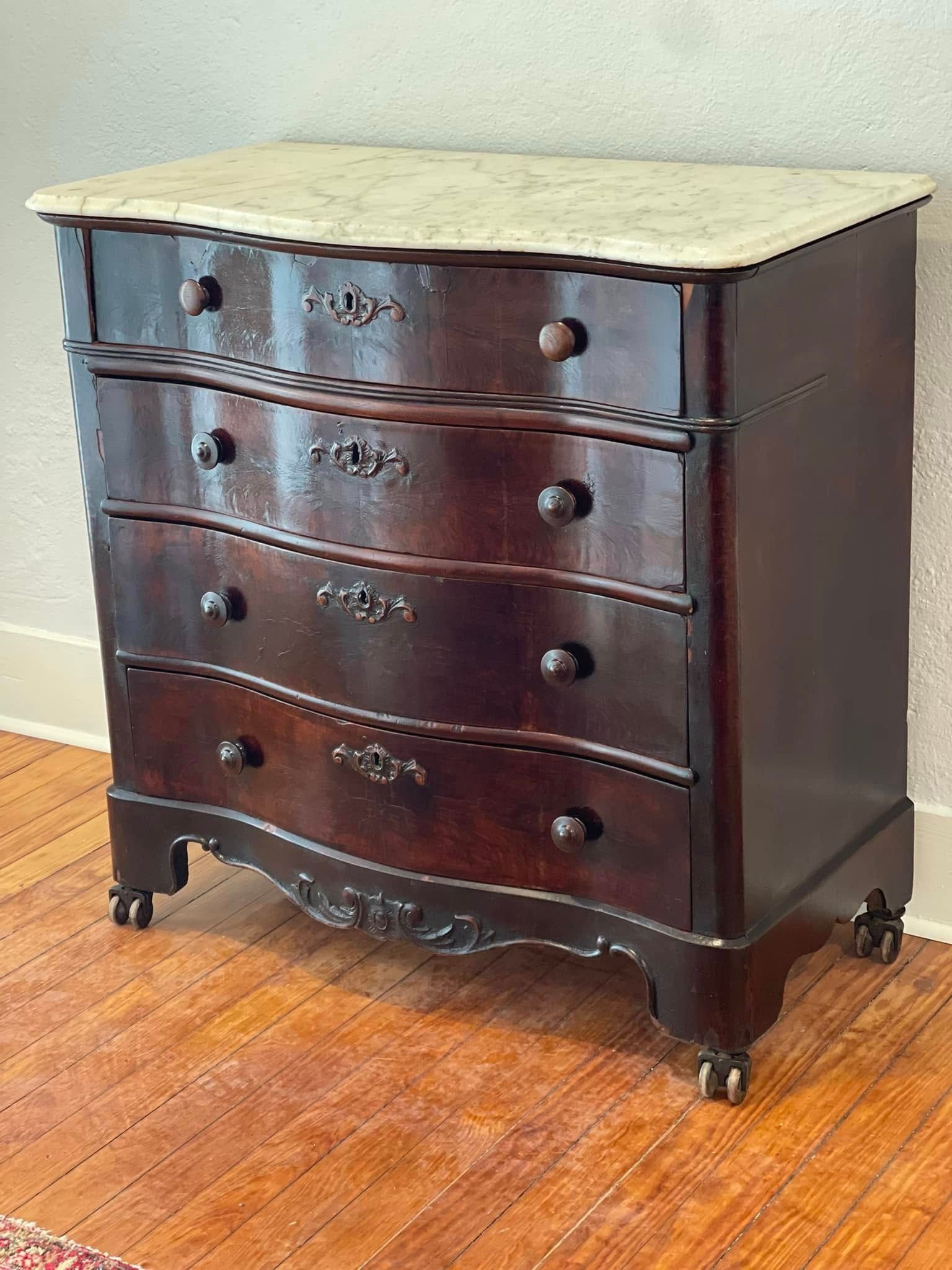 Dark wooden chest of drawers with a marble top, four drawers, and decorative hardware, set on a wood floor.