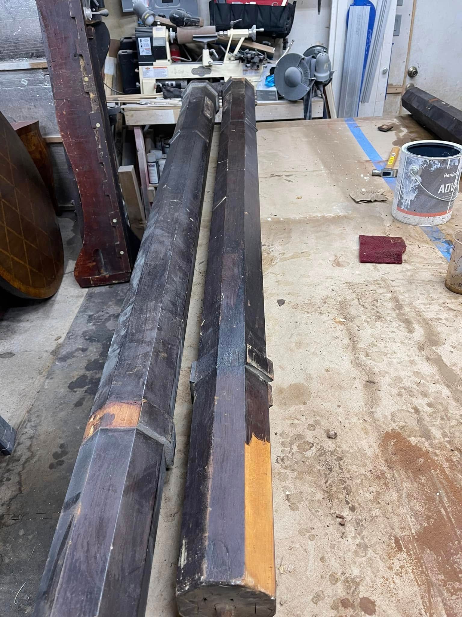 Two weathered, octagonal wooden posts lie in a workshop. A lathe and other tools are in the background.