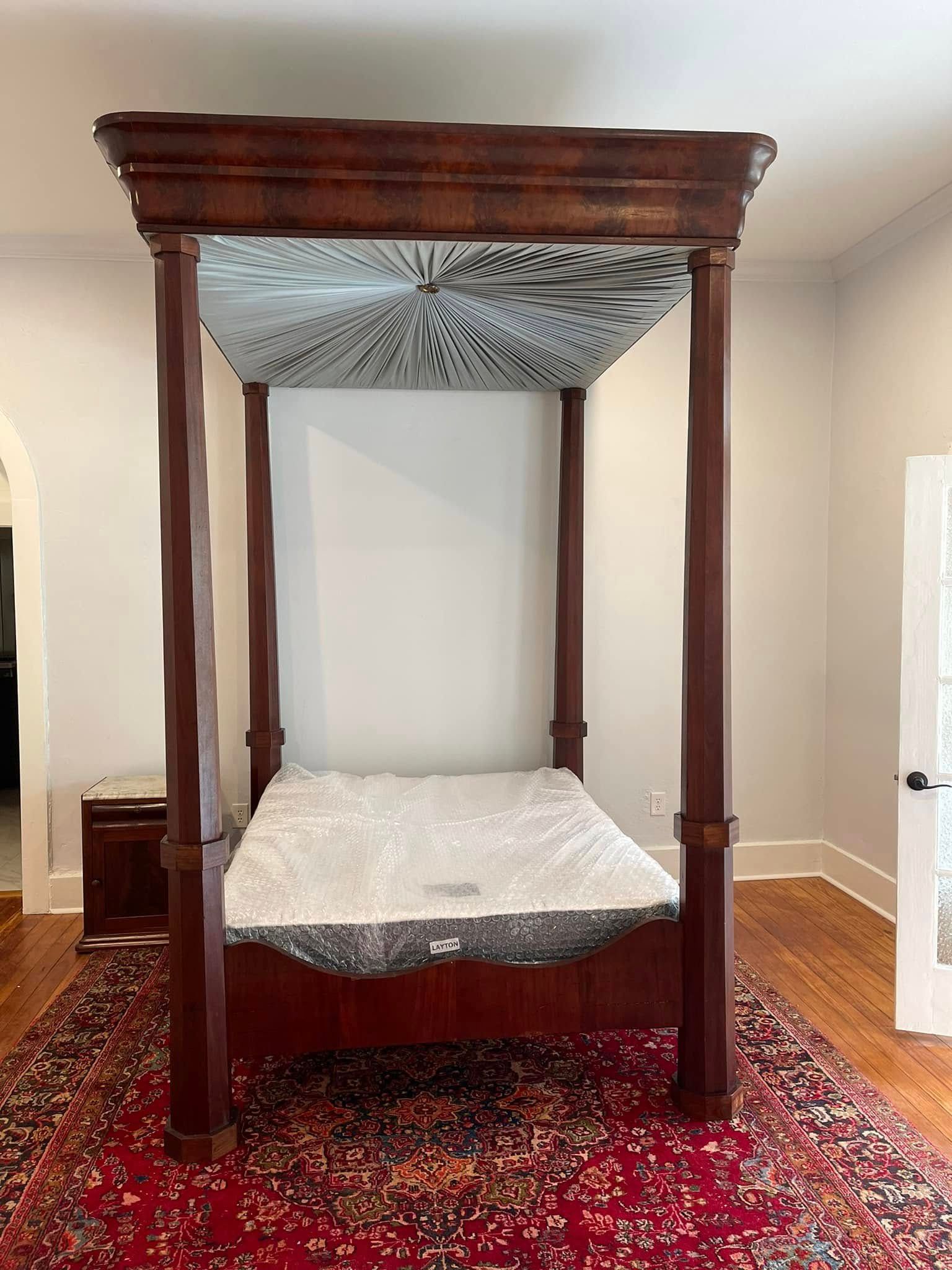 Four-poster bed with a mattress, set on a red rug in a room with hardwood floors.