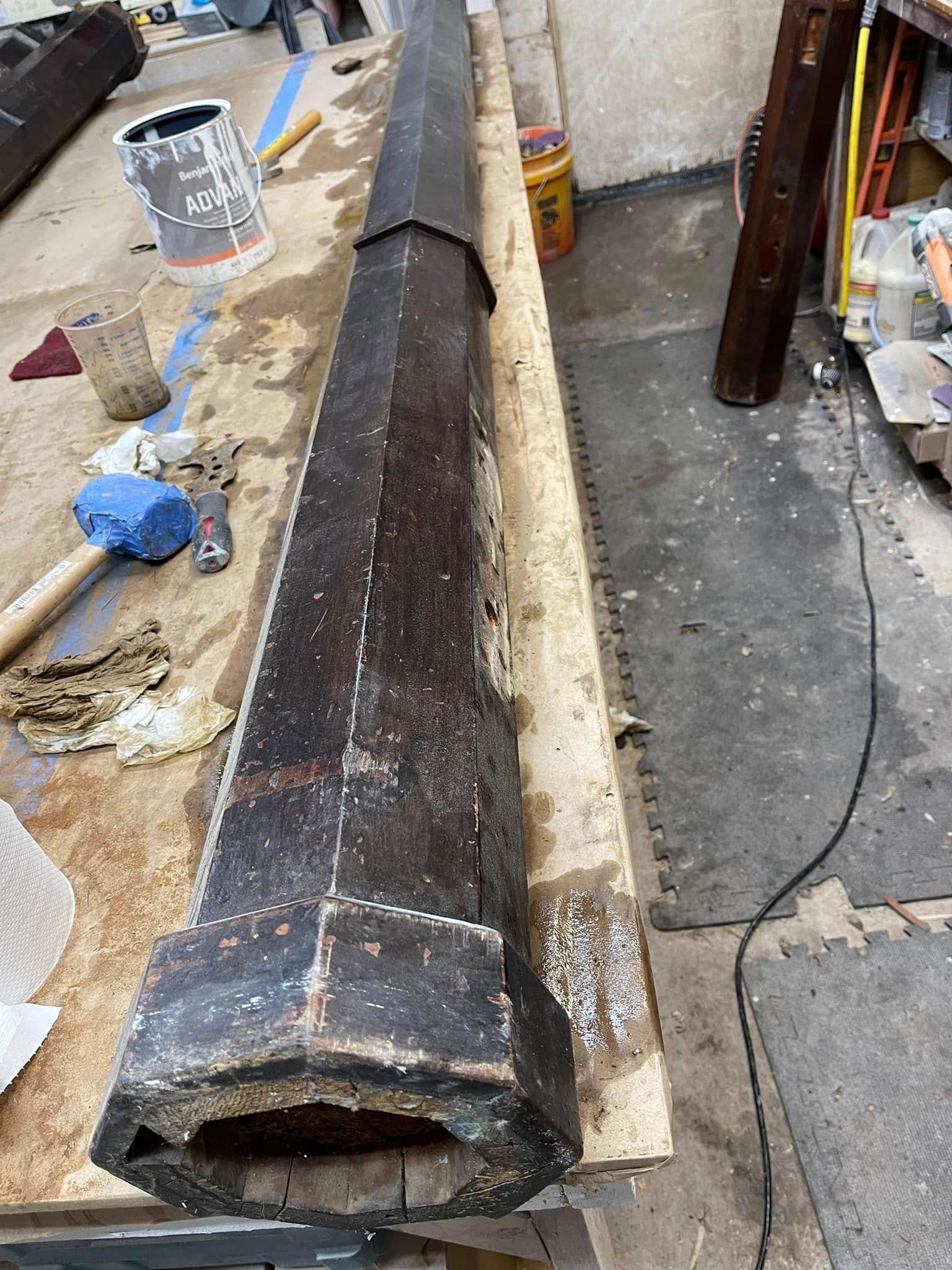 Long, dark, octagonal object with a wooden frame, lying on a work surface, tools in background.
