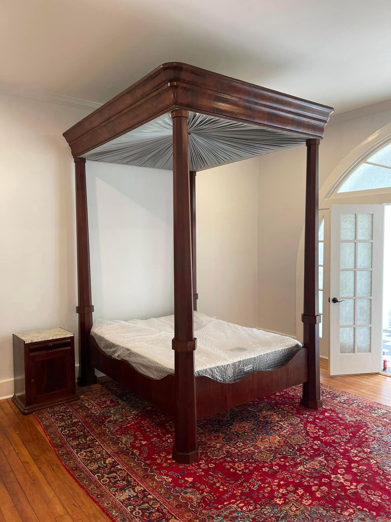 Four-poster bed with canopy, wood frame, sitting on a red oriental rug, next to a nightstand, in a room with a door.