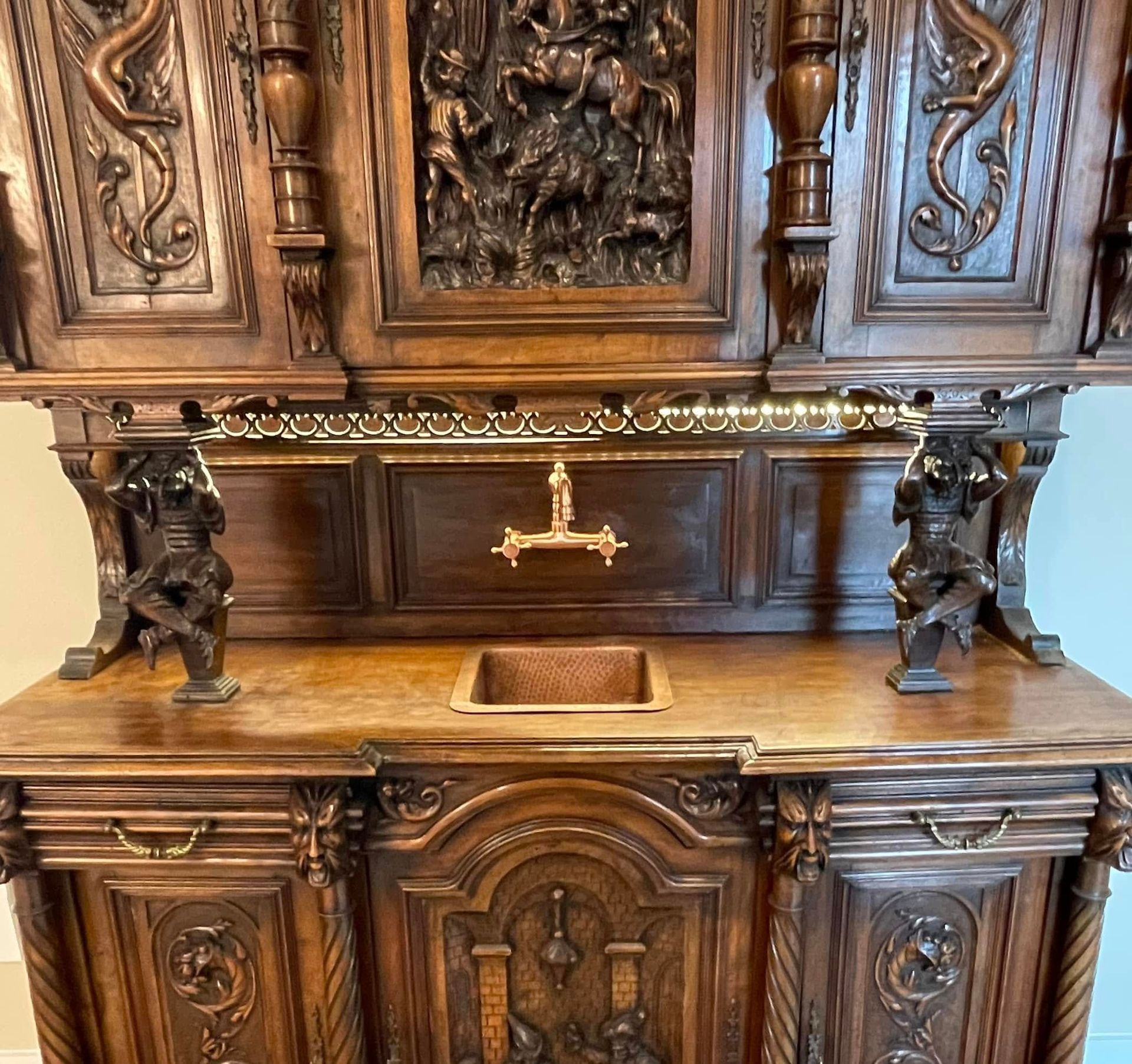 Ornate wooden cabinet with carved figures, featuring a sink and decorative top.