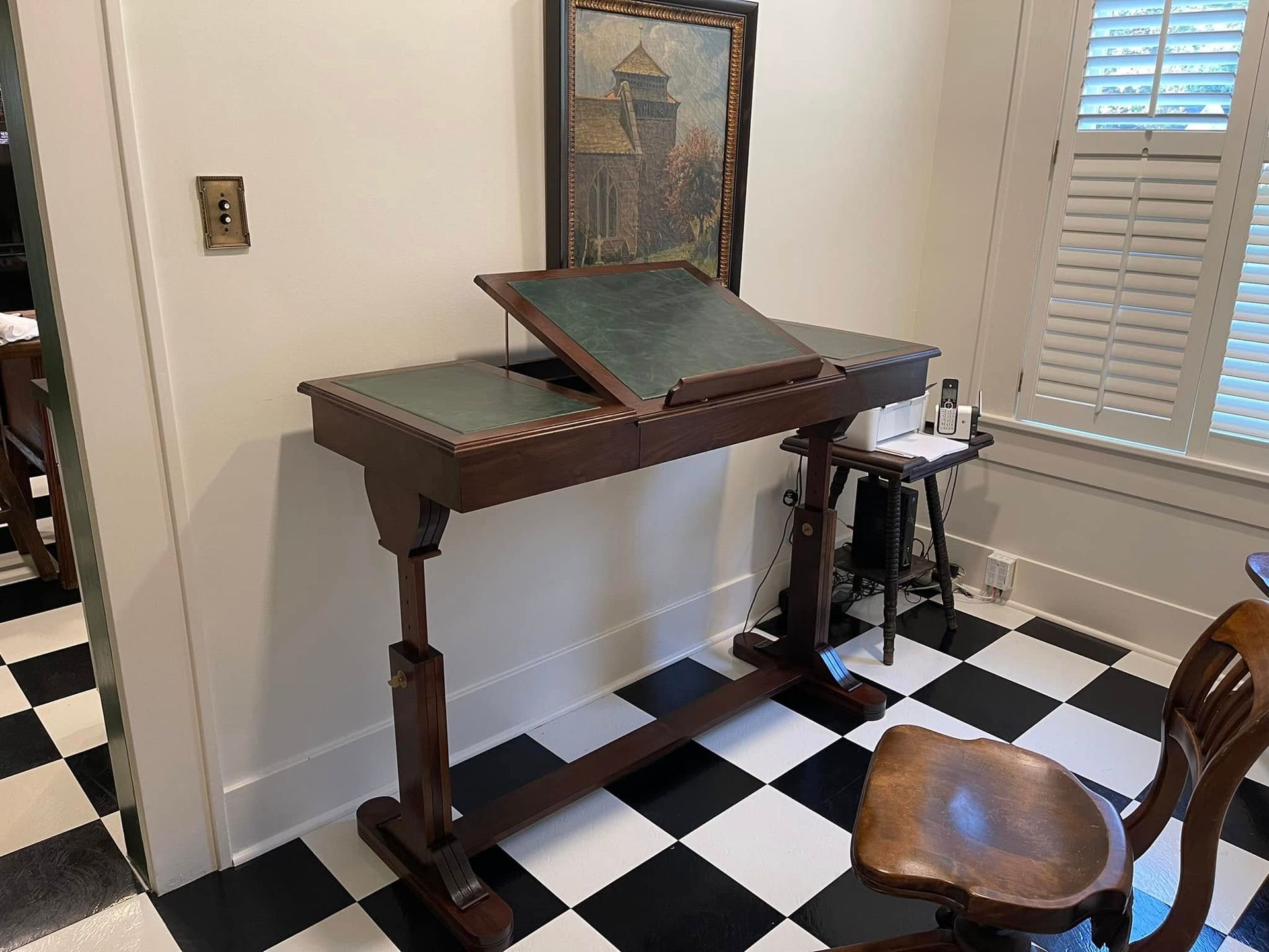 An antique wooden writing desk with a leather top, a painting, and a chair on a black and white tiled floor.