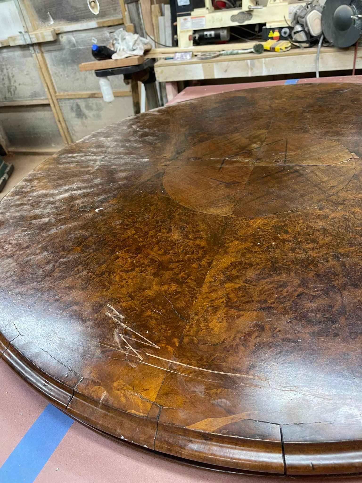 Round, wooden table top being restored in a workshop. Brown surface with scratches and white markings.