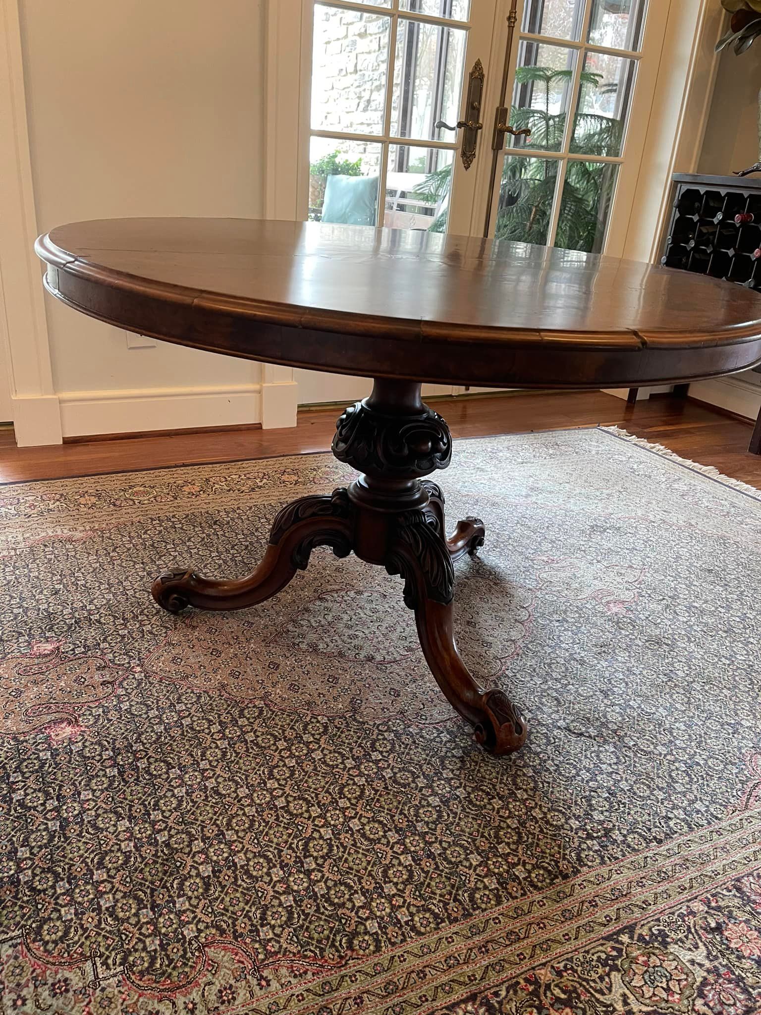 Round wooden table with ornate carved base on a rug, set in front of a door.