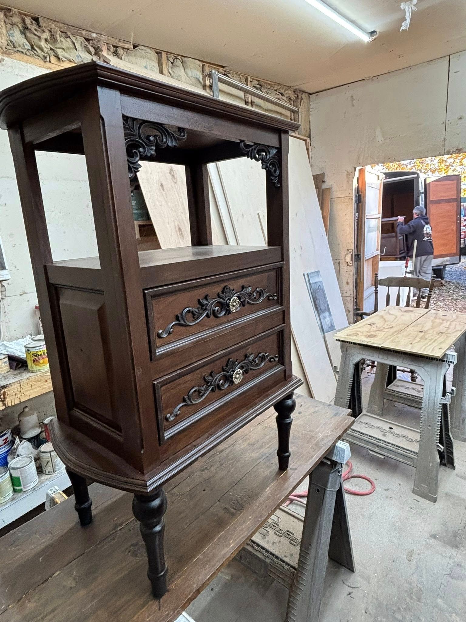 Dark wood antique cabinet with two drawers and open shelves, sitting on a wooden workbench.