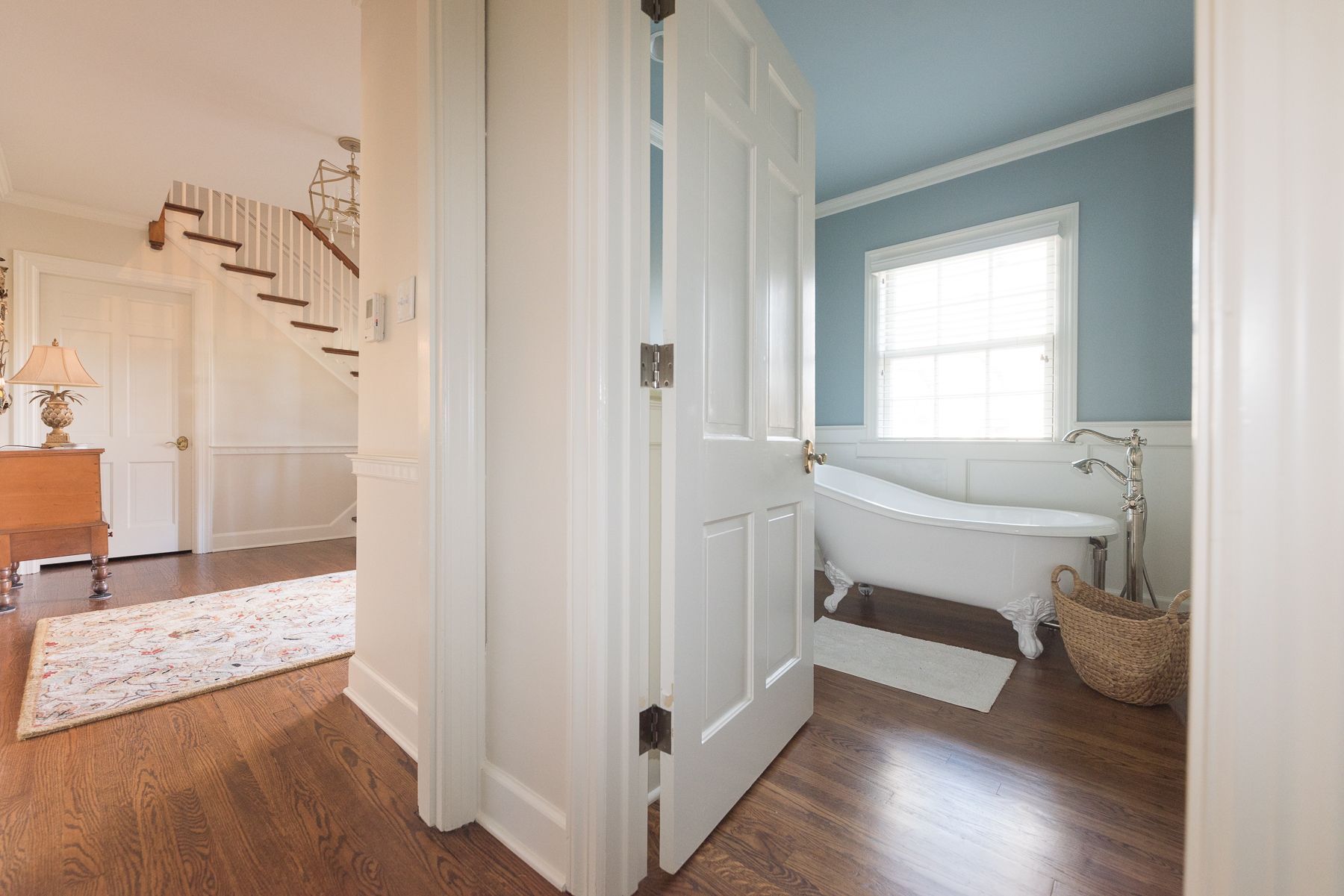 White doorway opens to a bathroom with clawfoot tub and hallway with staircase and wood floor.