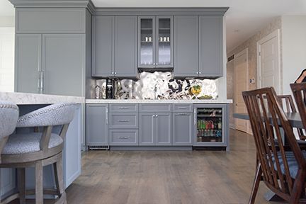 Gray cabinets in a kitchen with bar stools and dining chairs, a wine fridge, and a patterned backsplash.