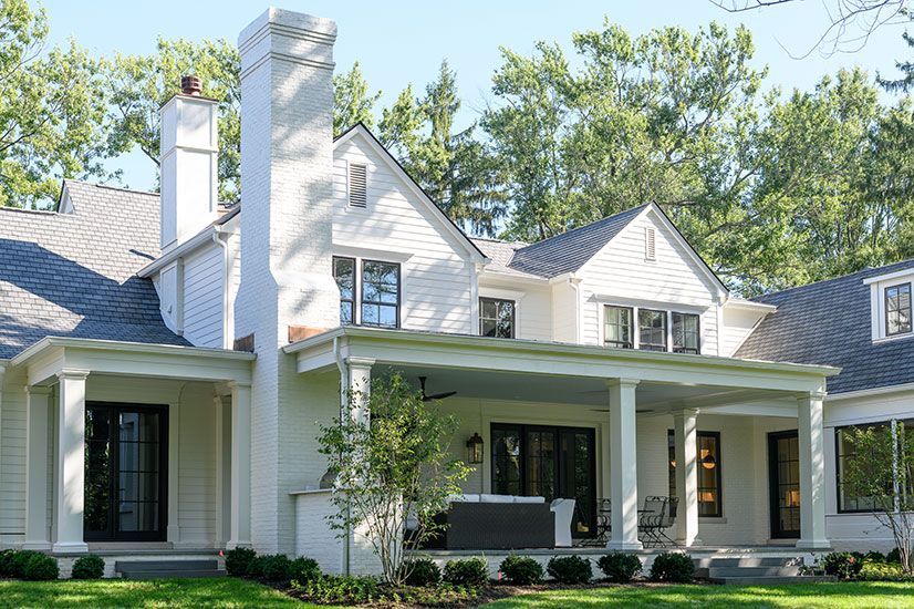 White two-story house with a porch, chimney, and black windows. Green trees and grass are in the background.