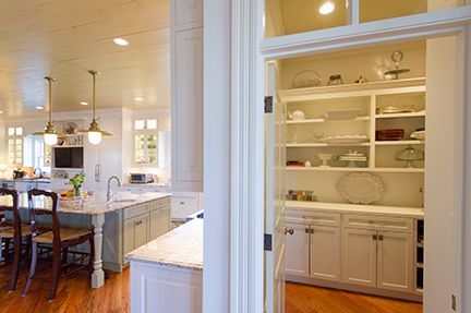 Kitchen with a pantry. White cabinets, marble counters, hardwood floors. Open door to a pantry with dishes on shelves.