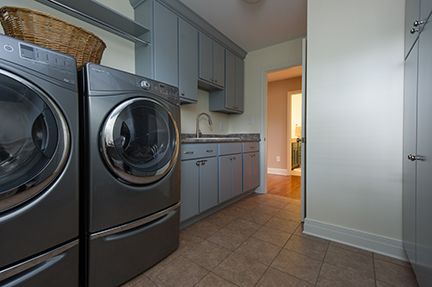 Laundry room with gray washer, dryer, cabinets, and sink. Beige floor tiles, basket, and doorway visible.