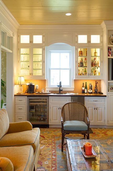 A cozy home bar with white cabinets, a wine fridge, and a window, lit with soft lighting.