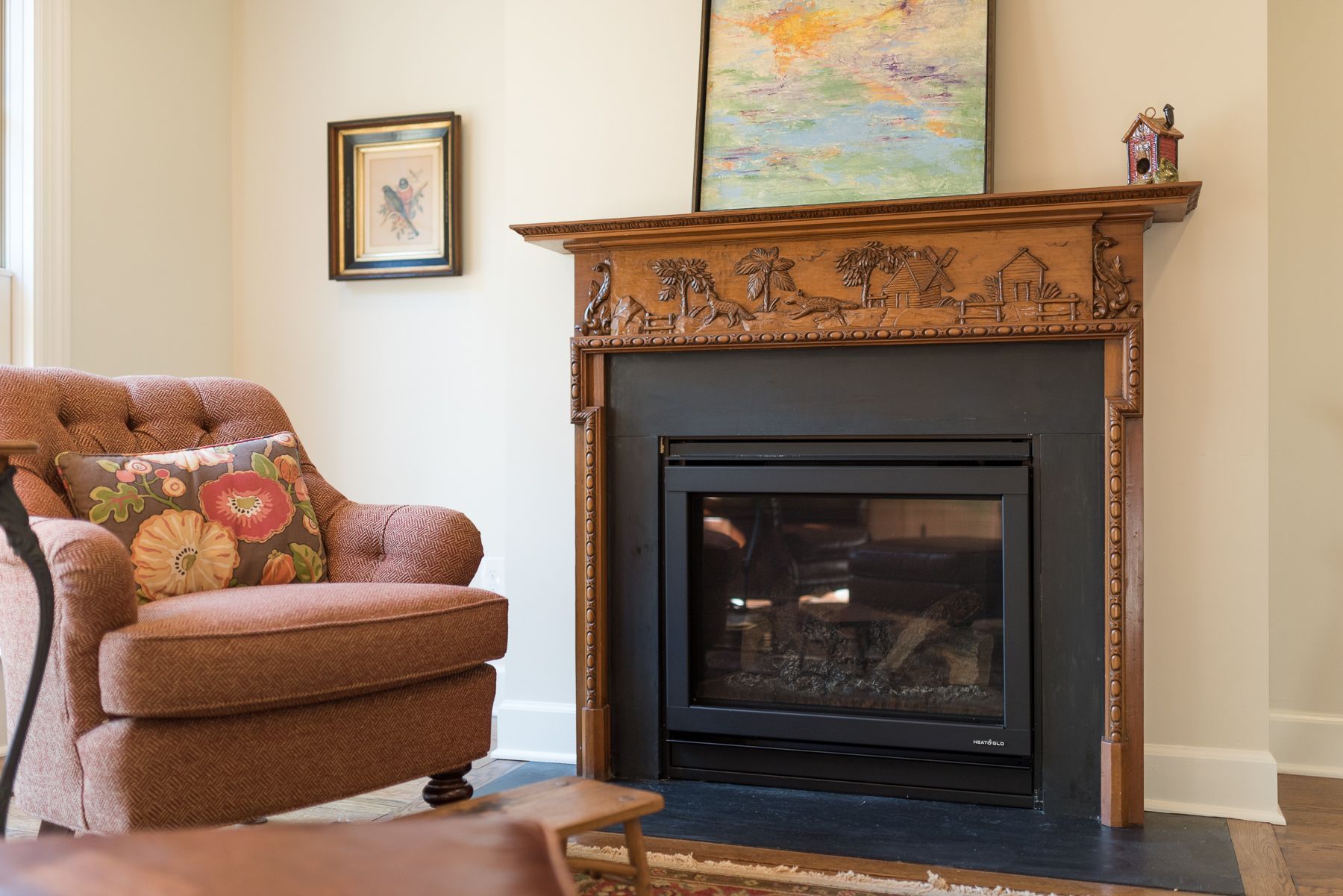 Cozy living room with brown chair and ornate fireplace, artwork on the wall, and a small wooden house on the mantel.