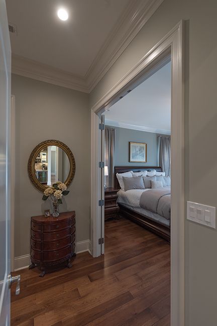 Hallway leads to bedroom with hardwood floors, ornate dresser, round mirror. Bedroom has bed with gray linens.