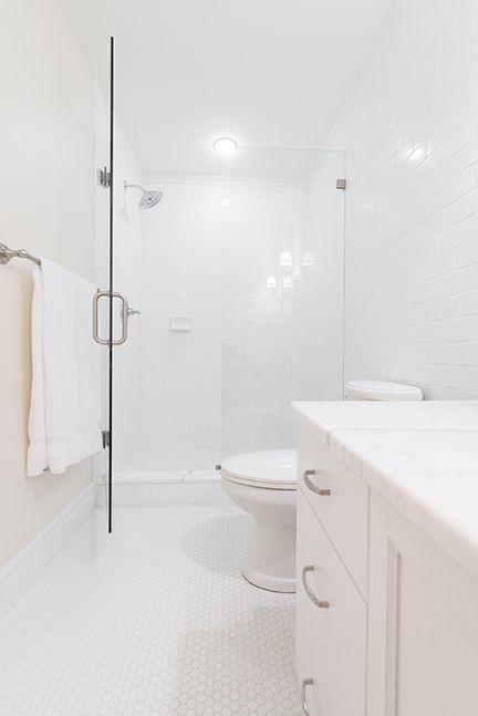 White bathroom with shower, toilet, and vanity. White tile floor and walls, glass shower door.