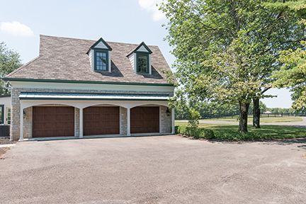 Three-bay garage with brown doors under a light blue-green trim and gray roof. Large tree to the right.