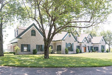 A light brick house with a green roof, shutters, and arched windows is framed by trees.