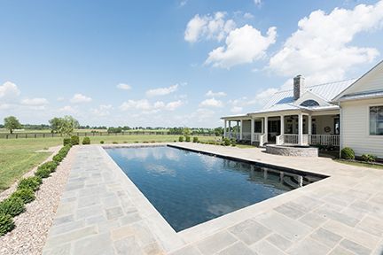 A rectangular pool next to a white house under a blue sky with a rural landscape in the background.