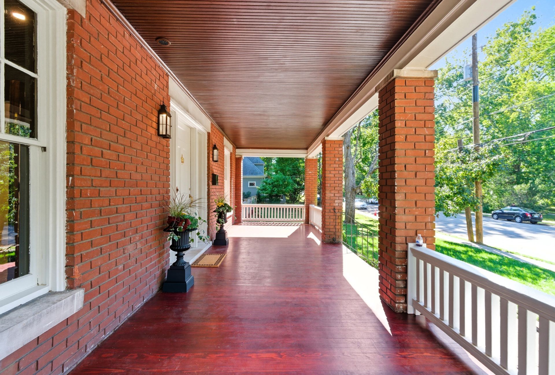 Long porch with brick walls, dark wooden ceiling, white railing, and potted plants.