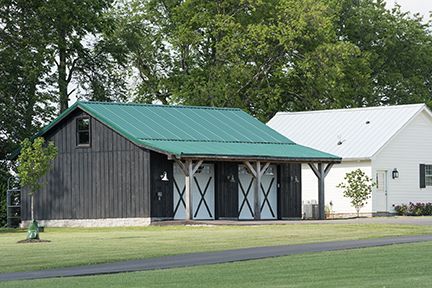 Barn with green roof and three stalls, next to a white building, on a grassy lawn.