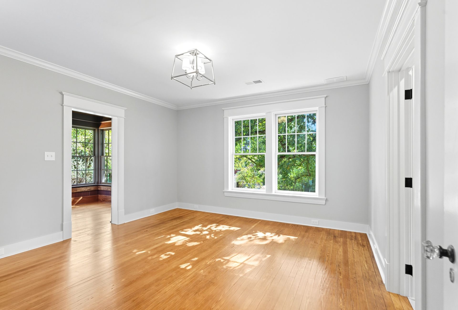 Empty room with hardwood floors, light gray walls, white trim, window with bright foliage view.