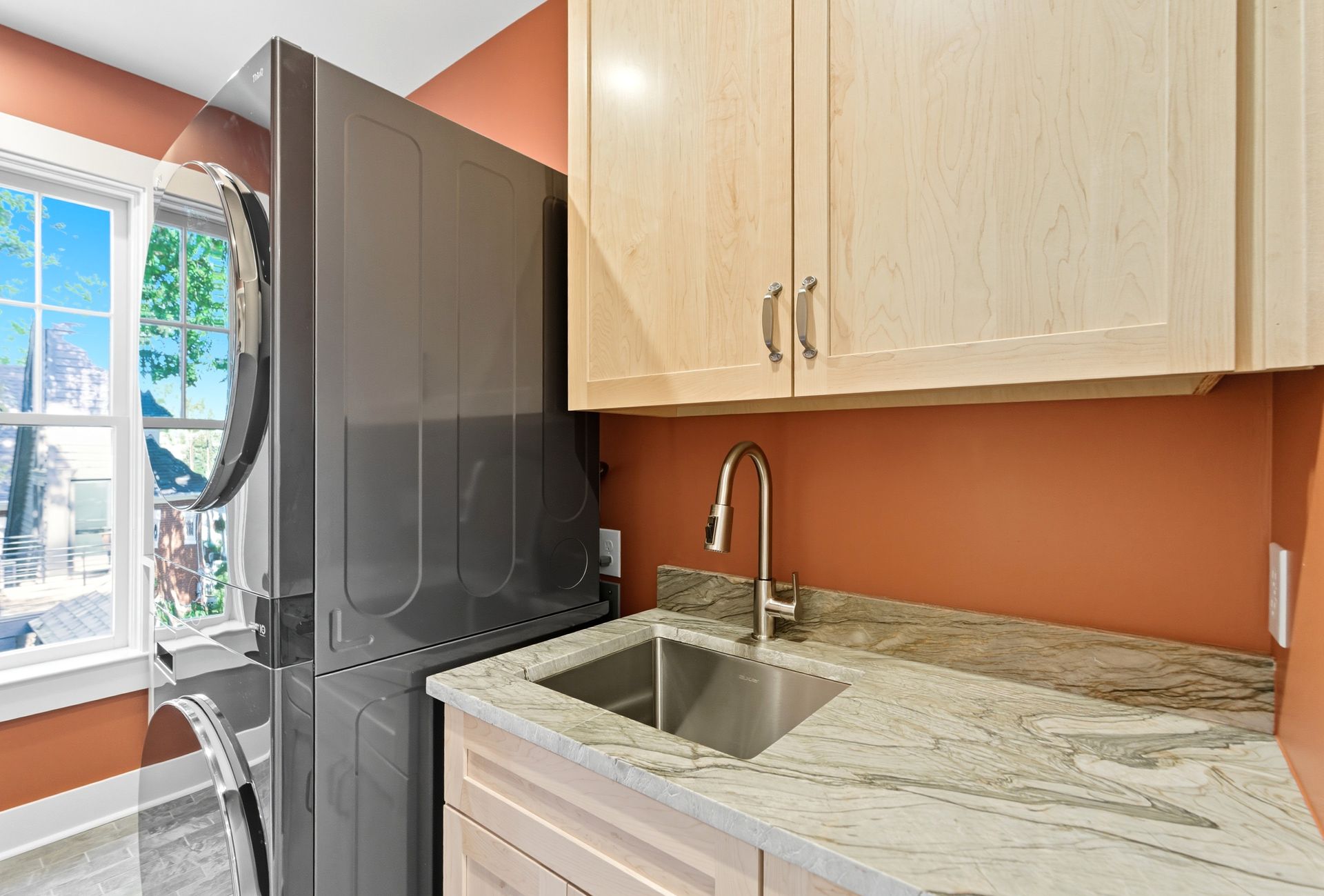 Laundry room with gray washer/dryer, sink, cabinets, window, and orange walls.