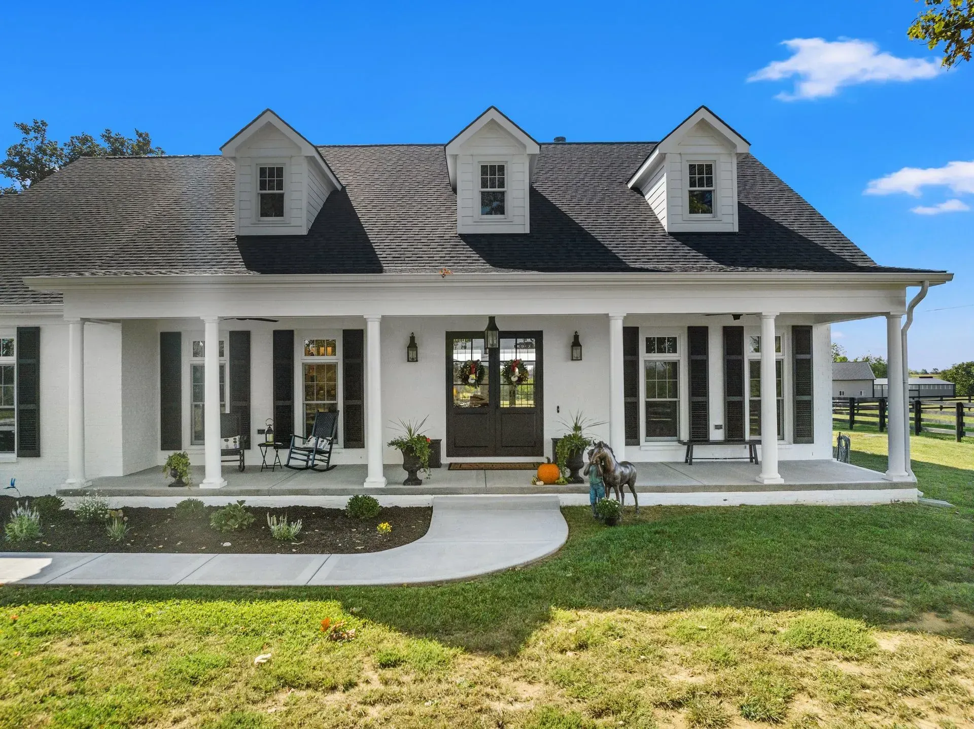 White farmhouse with black roof, dormers, and porch. Lush green lawn and blue sky.