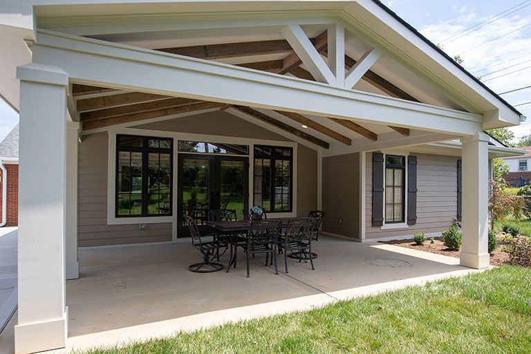 Beige covered patio with a black table and chairs, gray siding, and green grass.
