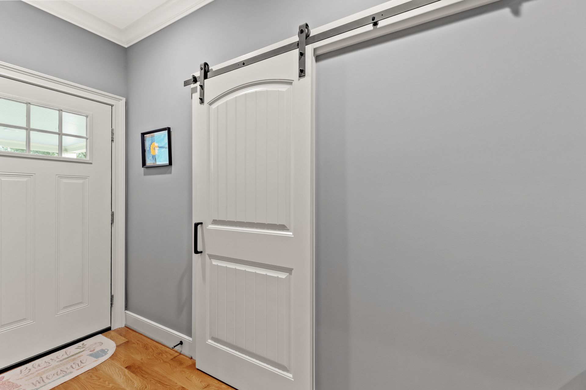 White barn door on a grey wall with black hardware. Wooden floor and white door to the left.