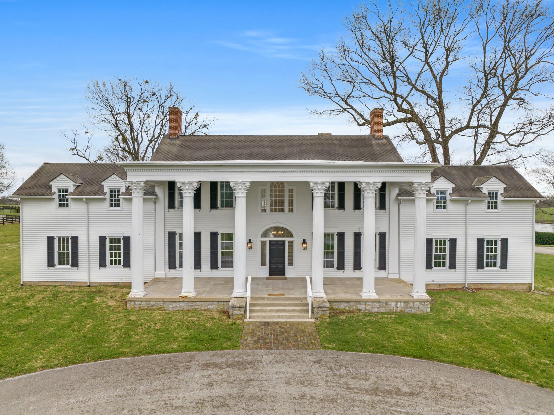 White two-story mansion with large columns and black shutters. Front door framed by pillars, set on a grassy hill.