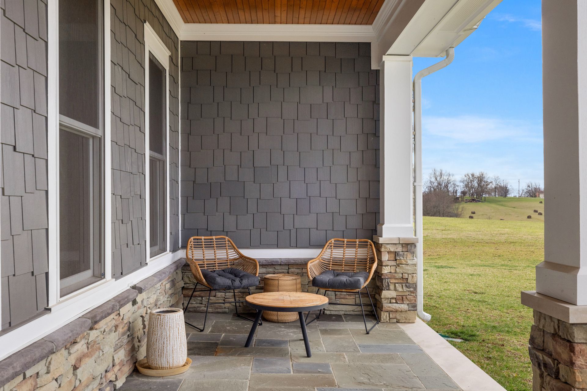 Two wicker chairs and small table on a porch with gray textured wall and stone base, open to a field.