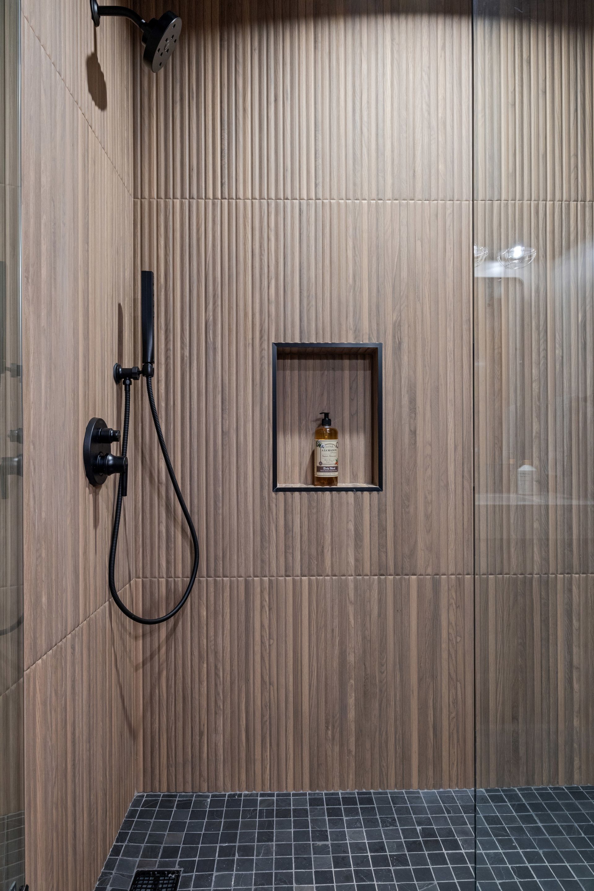 Shower with vertical, textured brown tile, black fixtures, and a recessed shelf holding a bottle. Dark tiled floor.