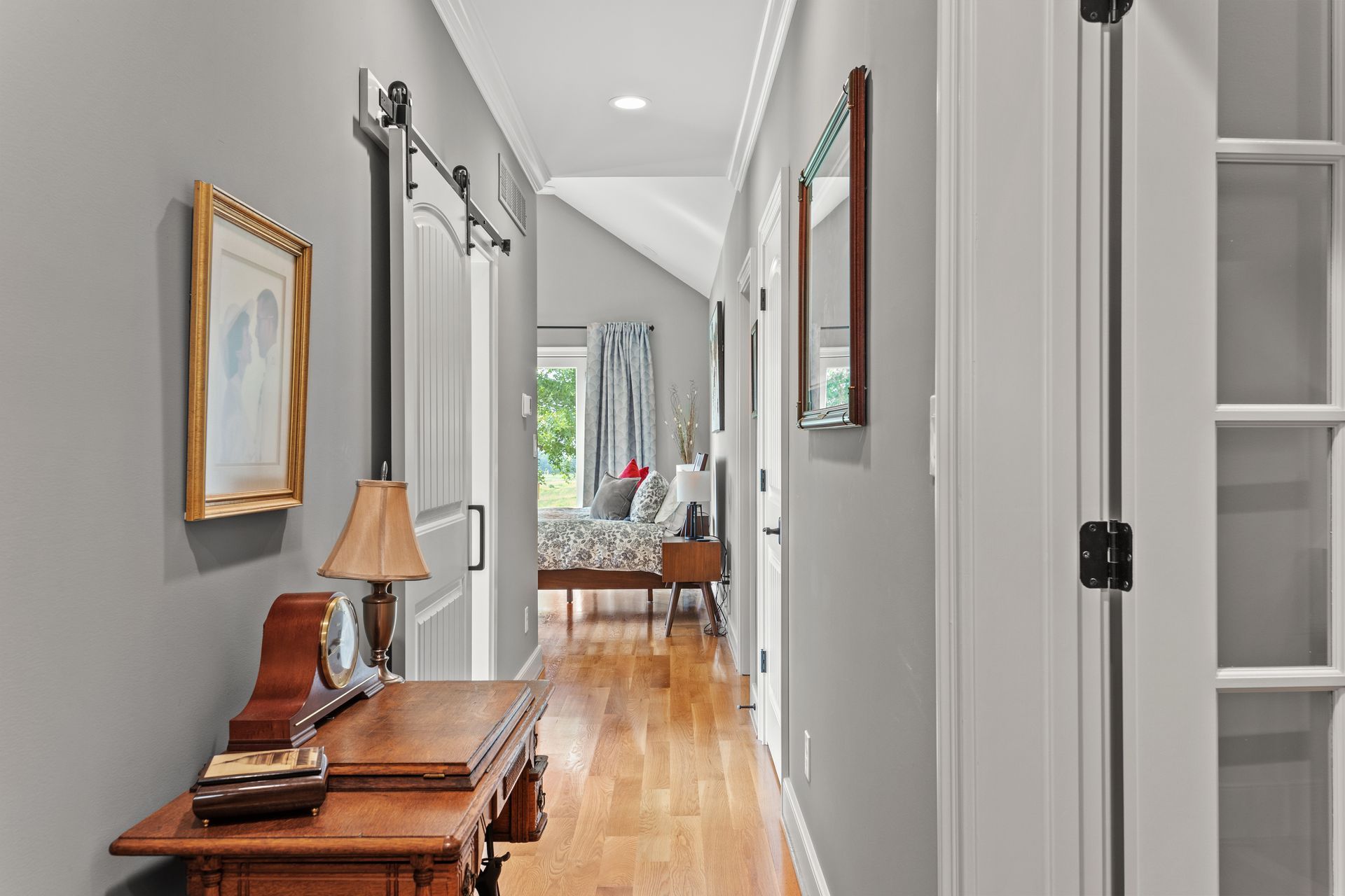 Long hallway with wooden floors, gray walls, and a view into a bedroom. Antique table and artwork present.