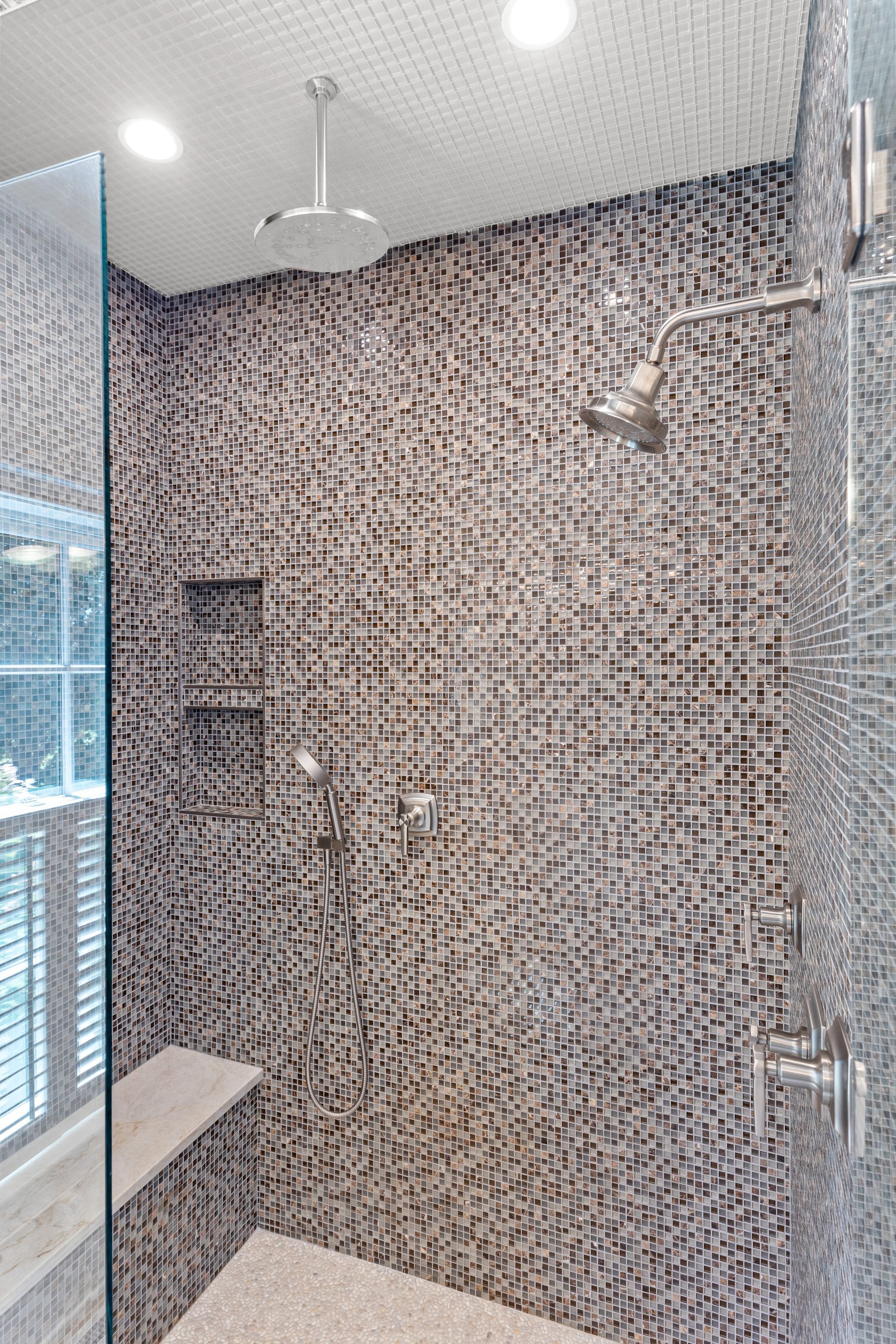 Shower with mosaic tile walls, glass door, built-in bench, and multiple shower heads.