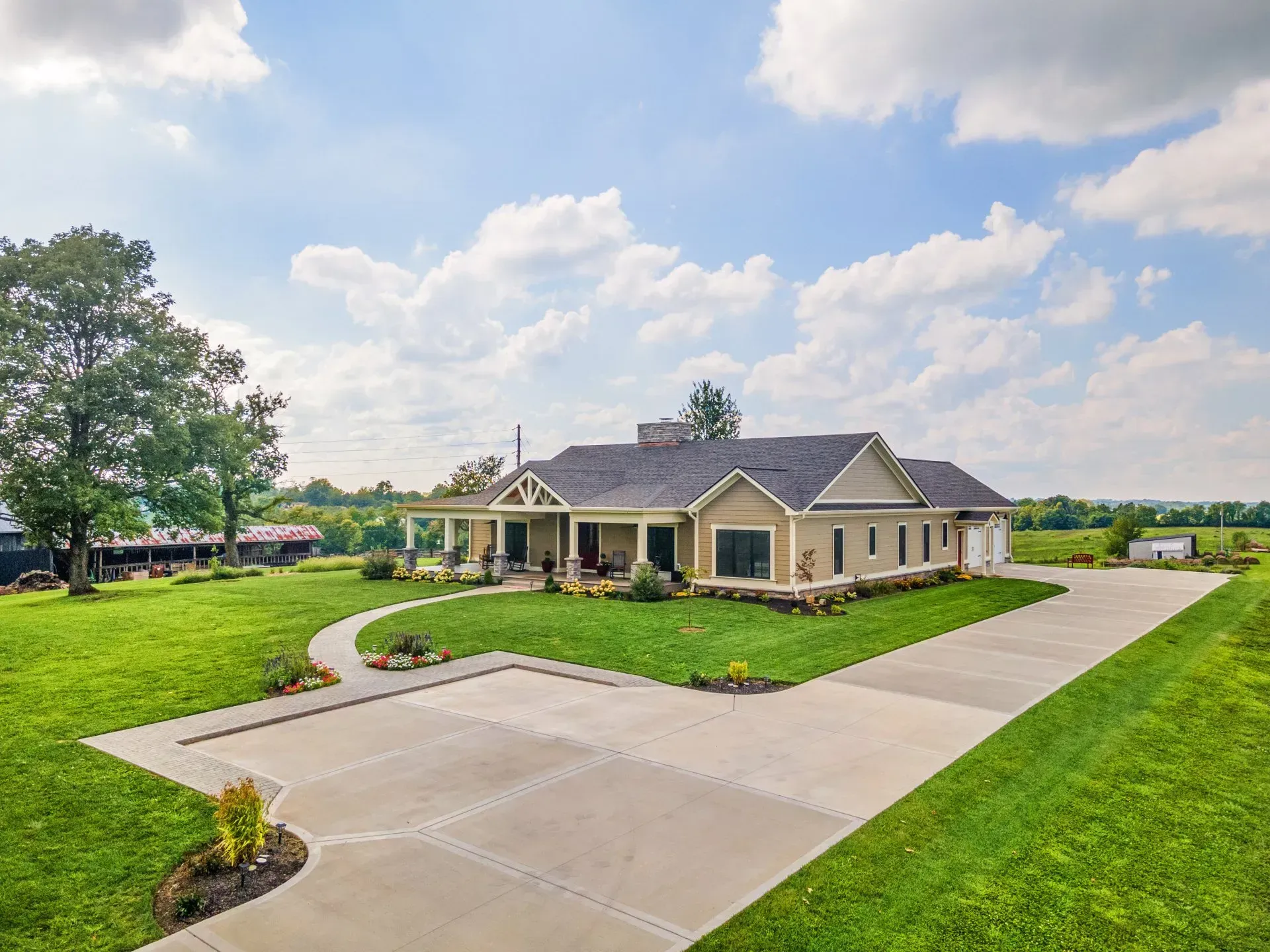 Beige farmhouse with long driveway on a grassy lawn under a partly cloudy sky.