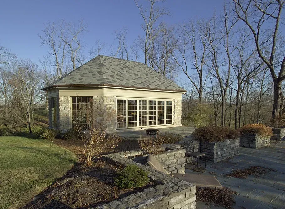 Stone gazebo with windows, set on a terraced stone patio and lawn, trees in the background under a blue sky.