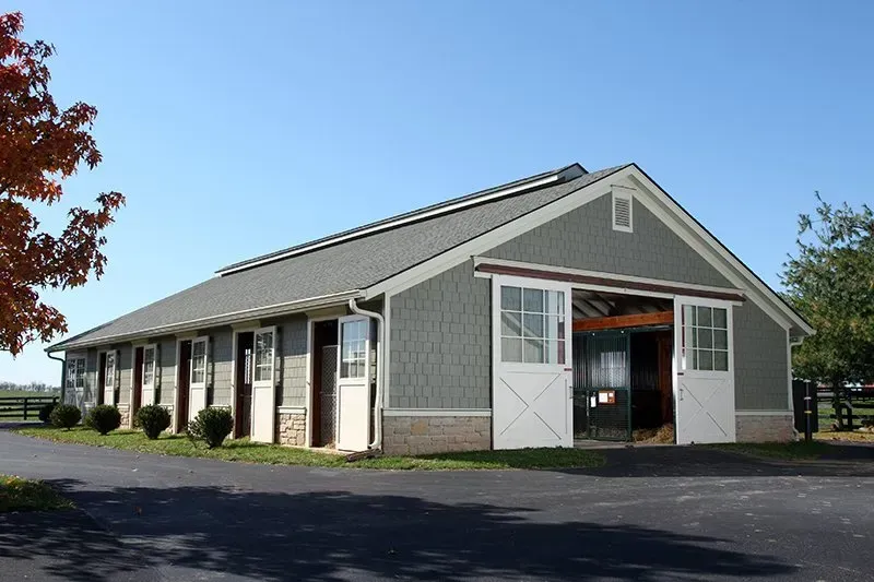 Horse barn with gray siding, white doors, and a black asphalt driveway.