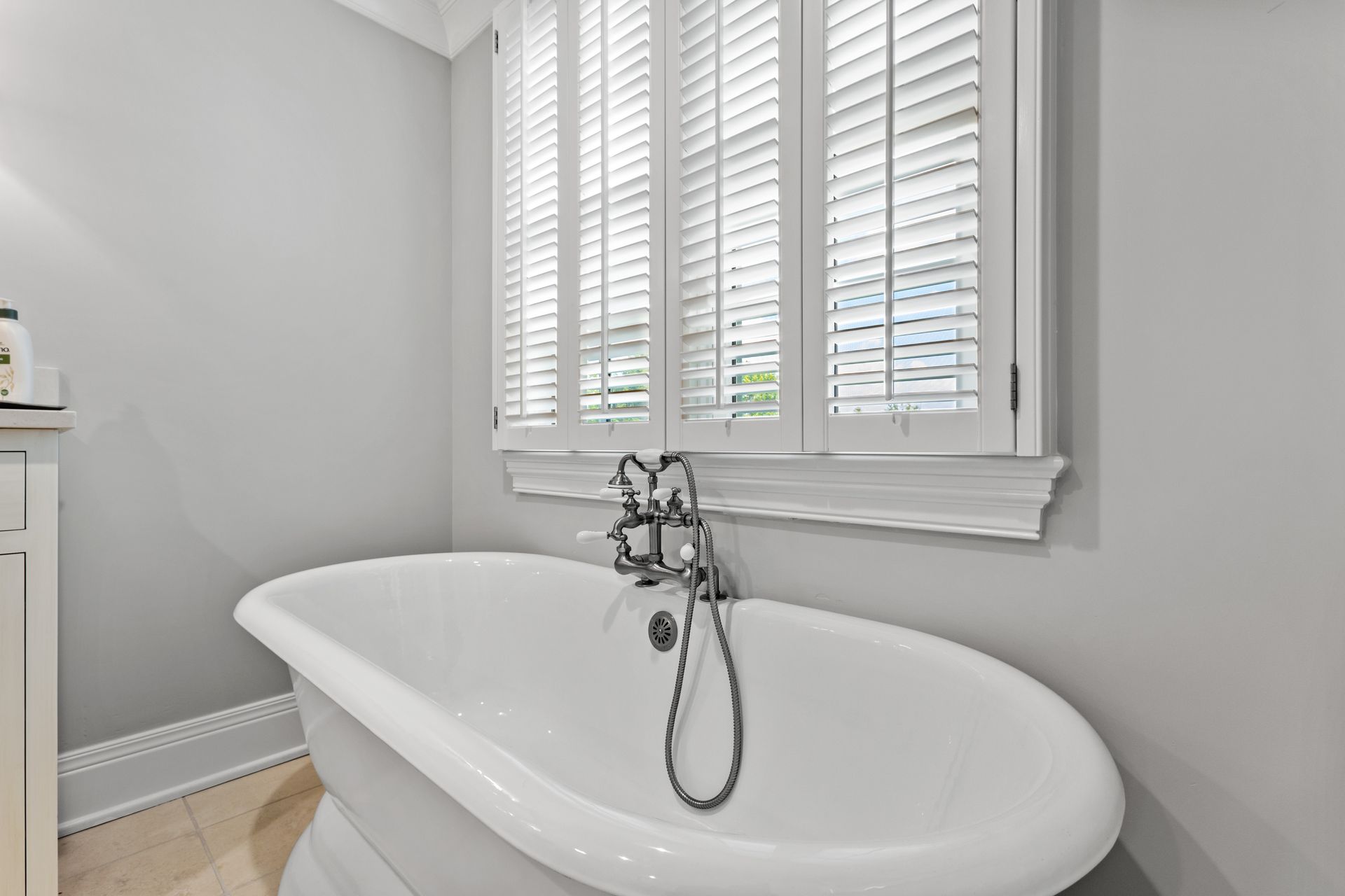 Claw-foot bathtub in a bathroom with gray walls, white window shutters, and light-colored flooring.