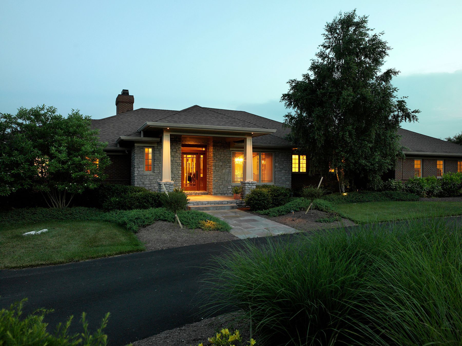 Stone house with a dark asphalt driveway, lit windows, and lush landscaping at dusk.