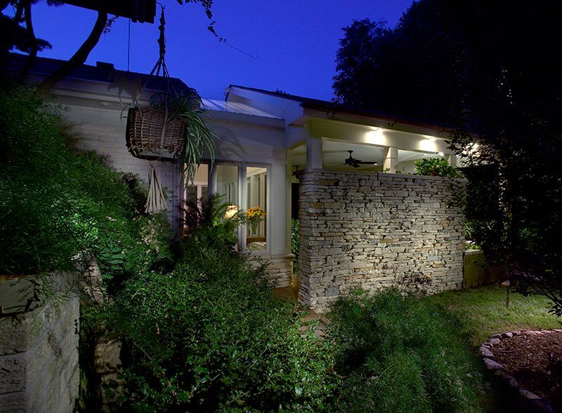 Nighttime view of a house with stone wall, porch, and lush greenery, illuminated by outdoor lighting.