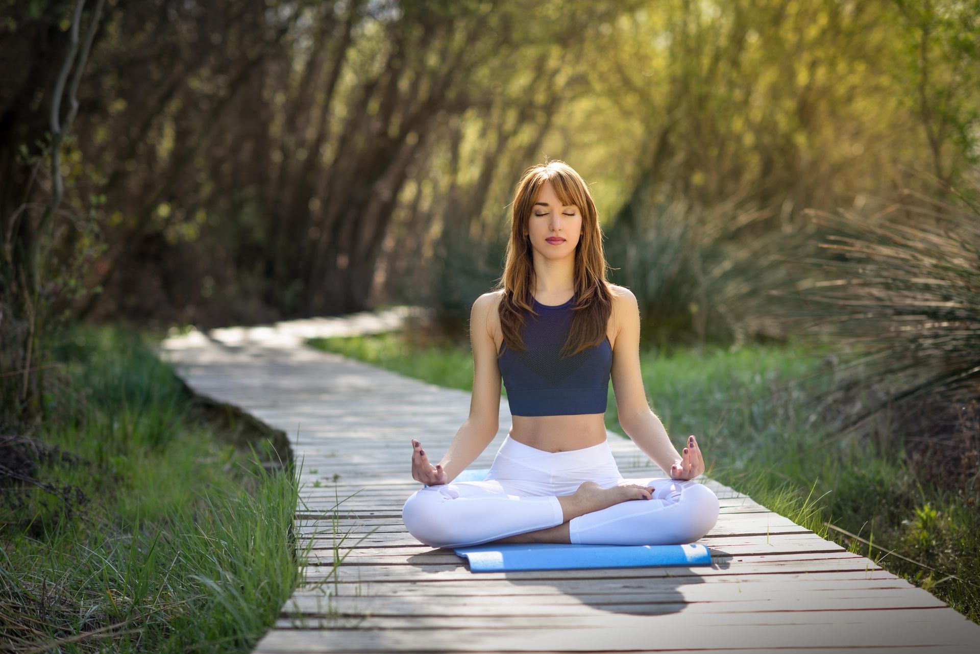 Person meditating on a blue mat on a wooden path in a green outdoor setting.