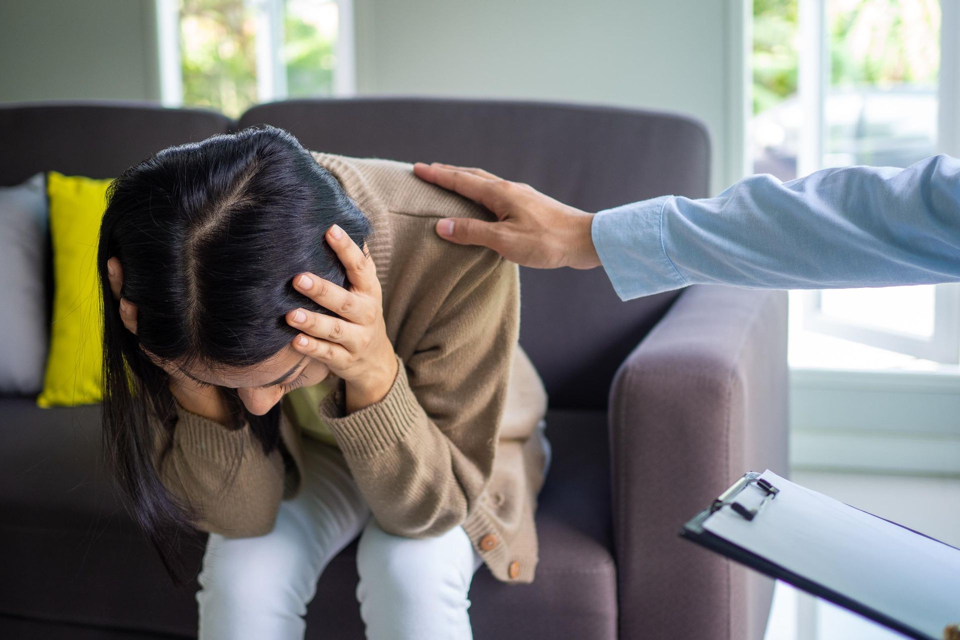 Distressed person on a couch with head in hands while another person comforts them with a hand on the shoulder