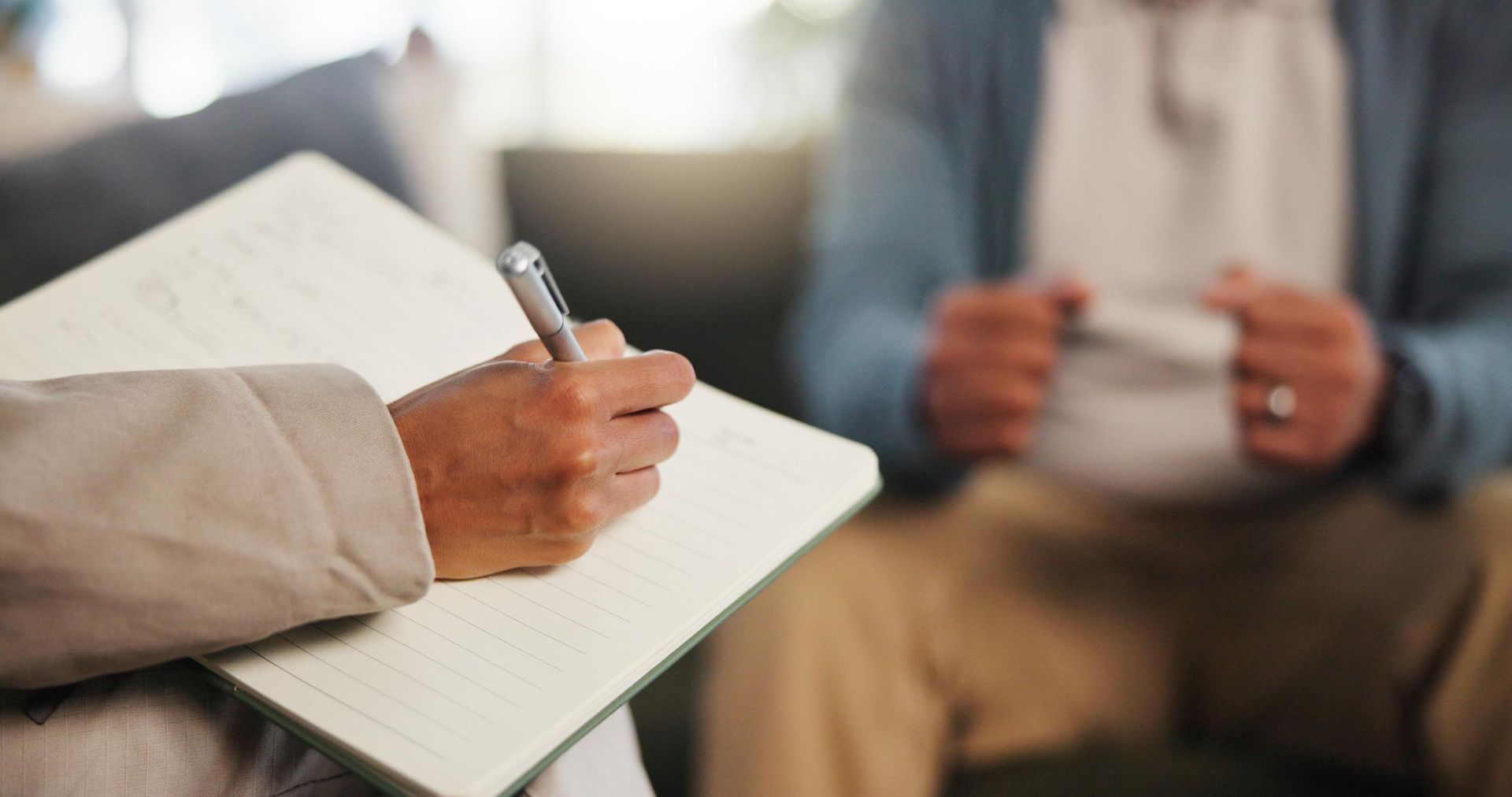 Person taking notes in a notebook while another sits across holding a cup.
