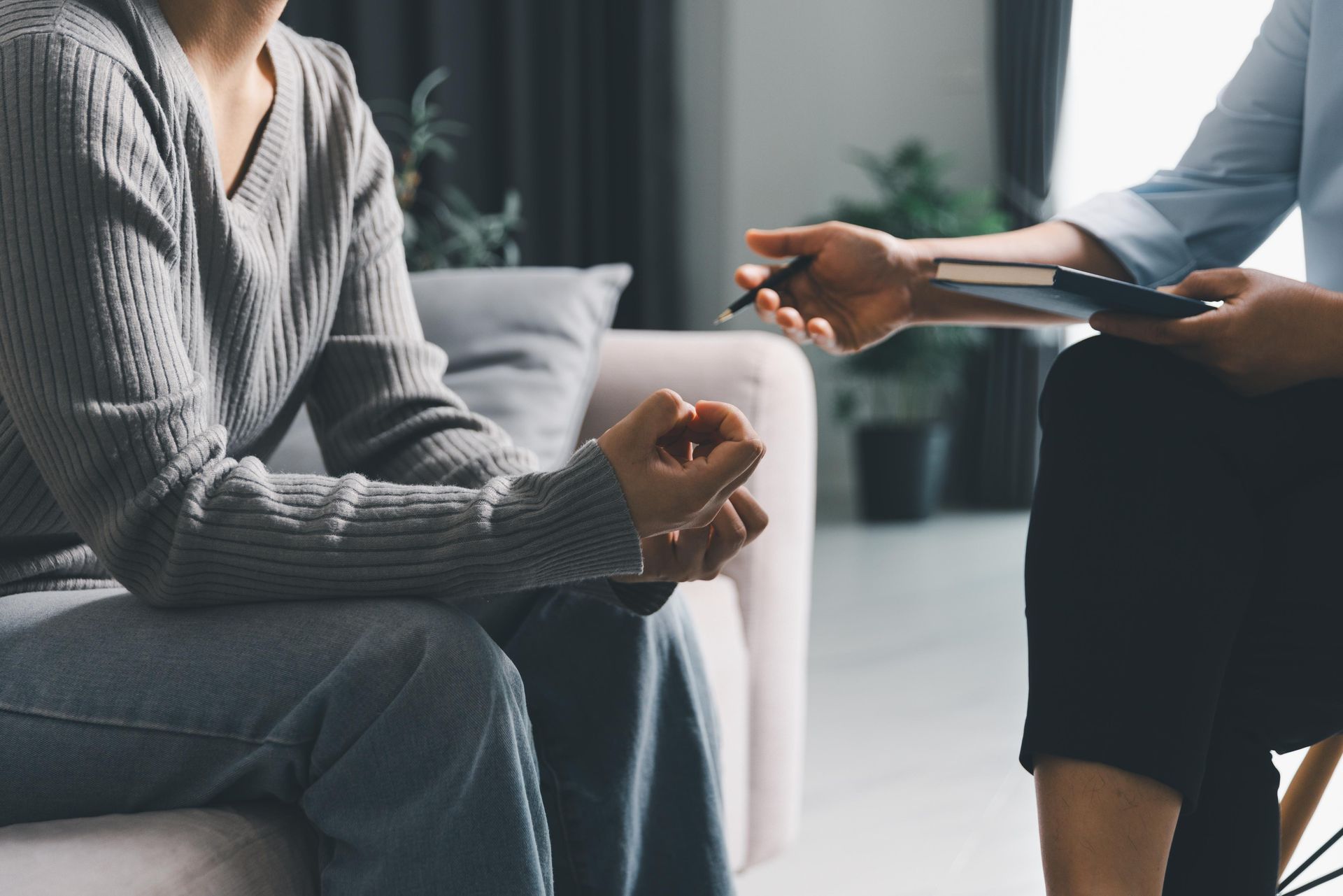 Two people seated in a counseling session, one holding a notebook and the other listening with folded hands
