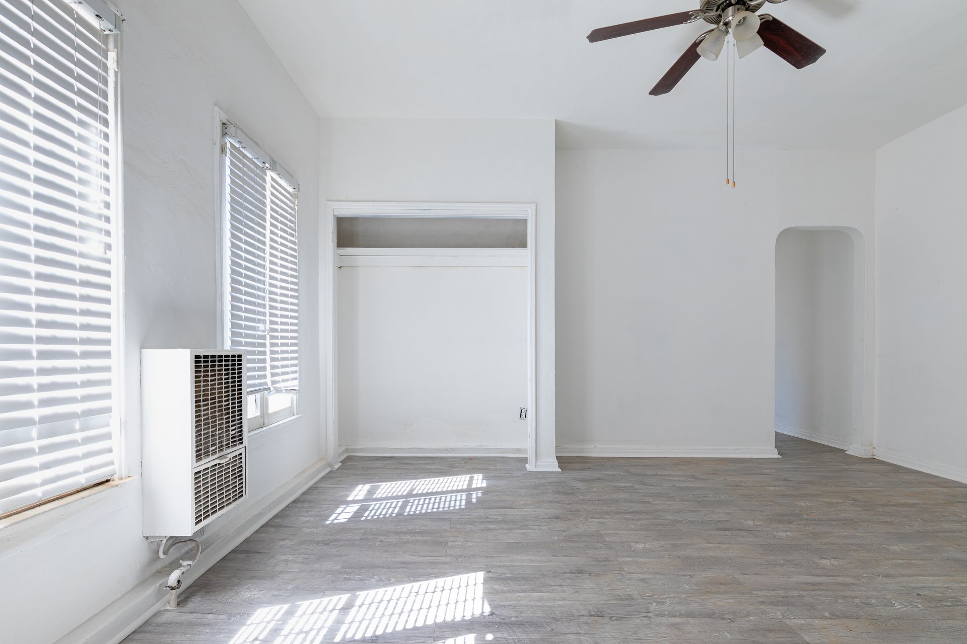 Empty, bright room with gray flooring, white walls, two windows with blinds, built-in storage, and ceiling fan.