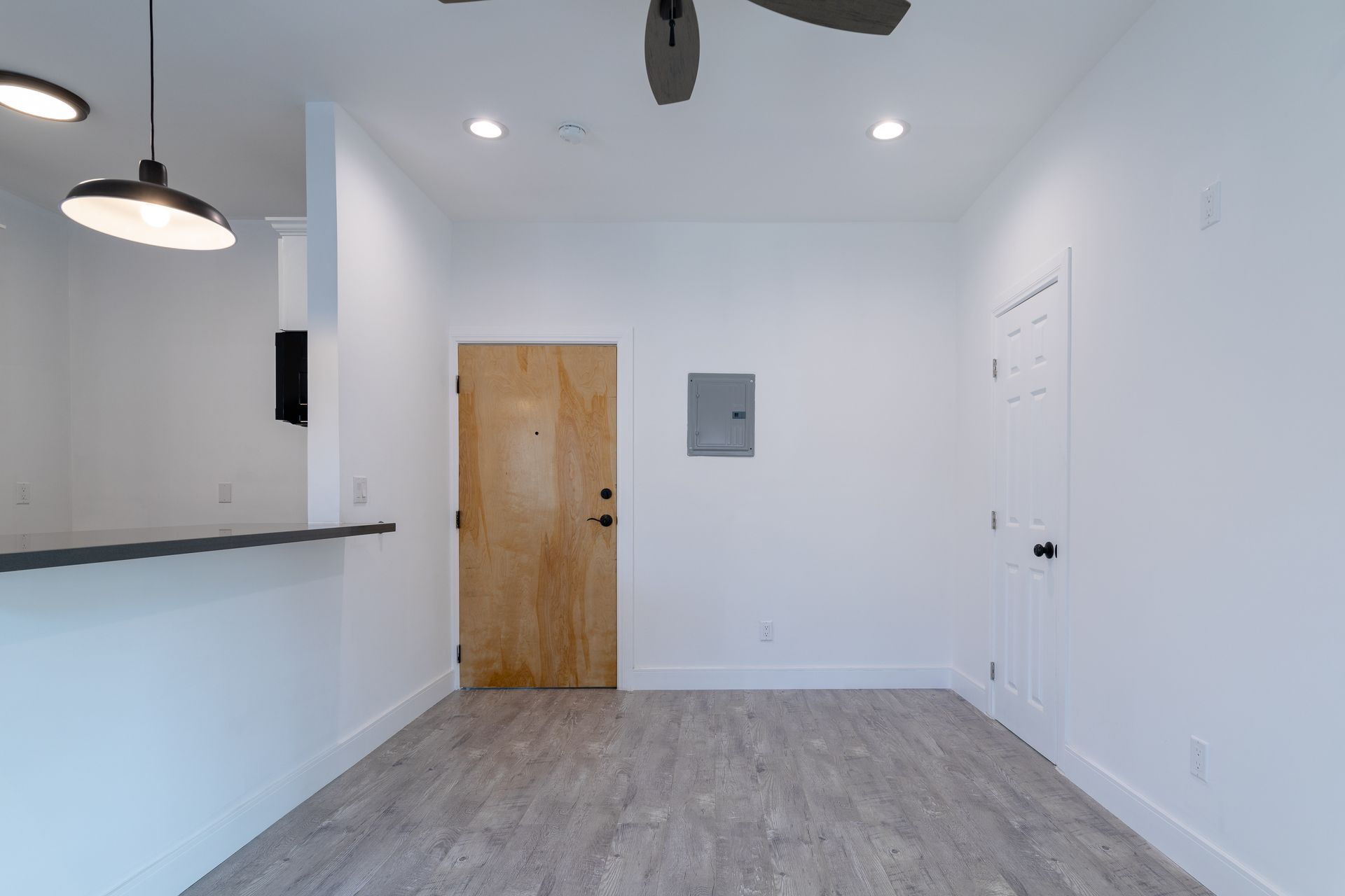 Interior of an empty, white-walled room with wooden door, gray floors, and a dark ceiling fan.