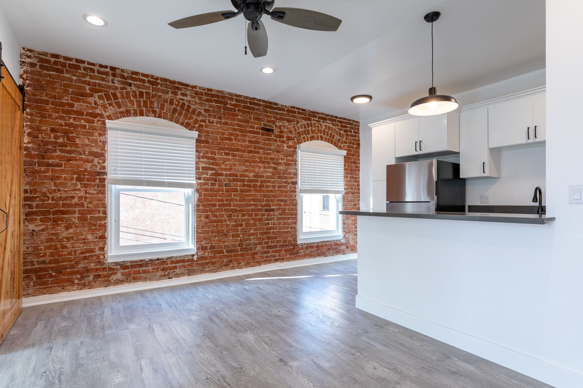 Living area with exposed brick wall, white kitchen, and ceiling fan.